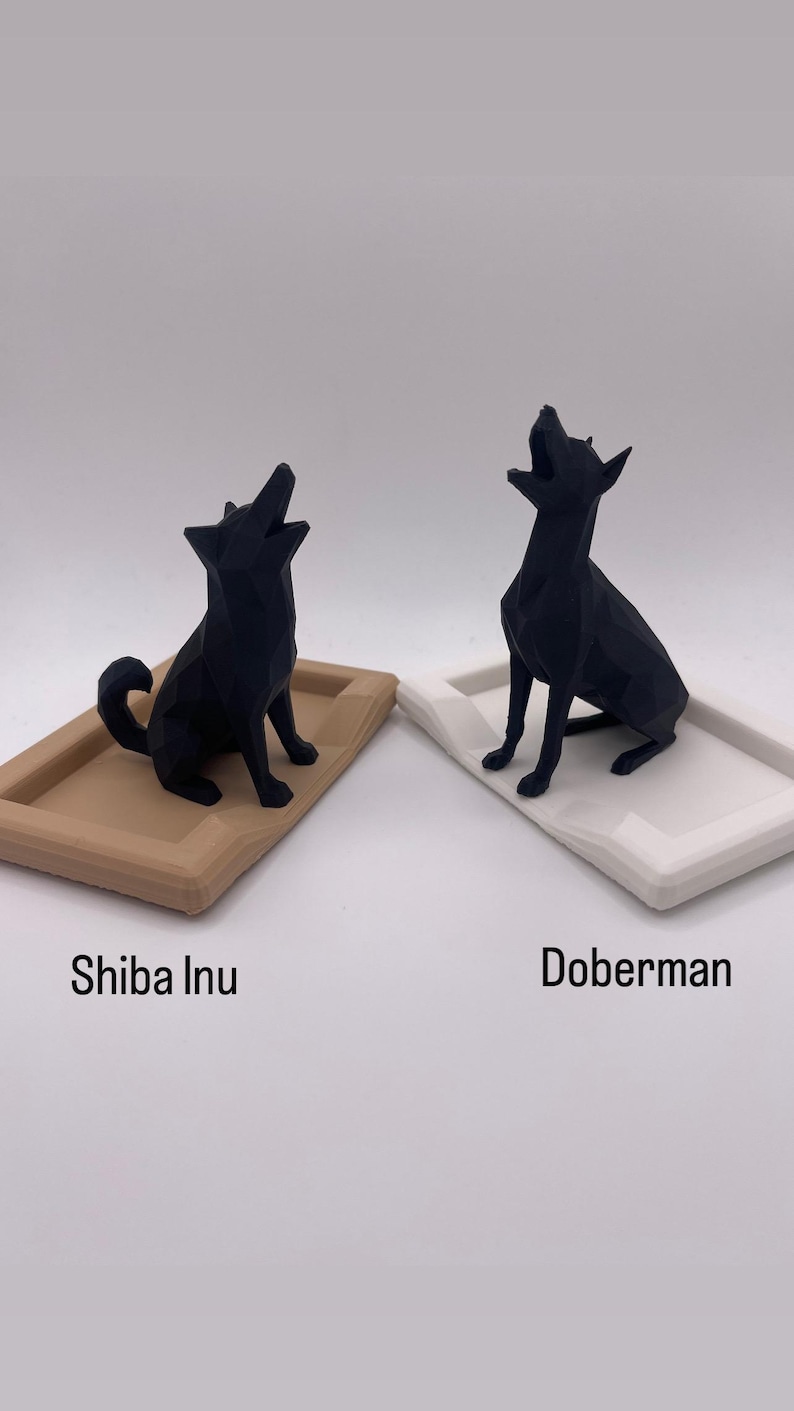 Puede incluir: Dos esculturas geom&eacute;tricas negras de perros, un Shiba Inu sobre una bandeja beige y un Doberman sobre una bandeja blanca. Ambos perros est&aacute;n en posici&oacute;n de aullido. Los textos "Shiba Inu" y "Doberman" est&aacute;n debajo.