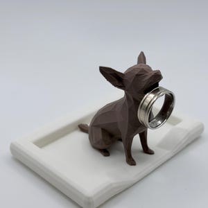 May include: A brown geometric chihuahua ornament holding a silver ring in its mouth, set on a white rectangular tray. The dog is the focal point, with a detailed, angular design. The ring is silver with an inscription.