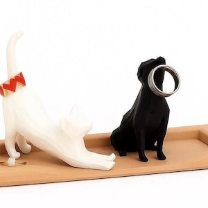 May include: A wooden tray with a white cat figurine stretching and a black dog figurine, both designed to hold rings. The cat has a red and gold collar. The dog holds a silver ring.