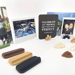 May include: A collection of rectangular photo stands in brown, tan, and black, alongside small decorative shapes resembling clouds and crystals. A black square sign displays the white text: CELEBRATE ANY PROGRESS. DON'T WAIT TO GET PERFECT. Ann McGee-Cooper.
