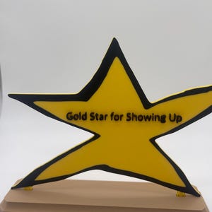 May include: A yellow star-shaped plaque with a black outline and the black text "Gold Star for Showing Up" printed across the center. The star is mounted on a rectangular, light brown base with beveled edges, creating a freestanding display piece.
