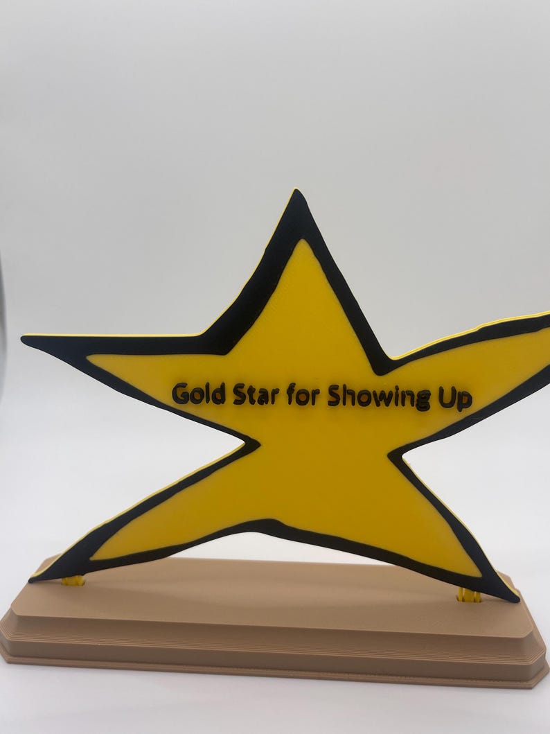 May include: A yellow star-shaped plaque with a black outline and the black text "Gold Star for Showing Up" printed across the center. The star is mounted on a rectangular, light brown base with beveled edges, creating a freestanding display piece.