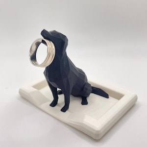 Puede incluir: Un soporte para anillos con forma de perro geom&eacute;trico negro con un anillo plateado en la boca. El perro est&aacute; sobre una bandeja rectangular blanca. El soporte para anillos es un objeto decorativo para guardar anillos y joyas.