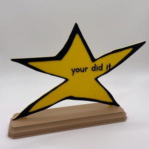 May include: A yellow star-shaped plaque with a thick black outline, featuring the black text "your did it" printed in the center. The star is mounted on a rectangular, light brown base with beveled edges, standing upright against a plain background.