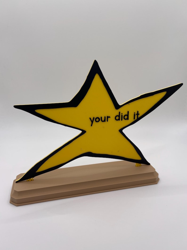 May include: A yellow star-shaped plaque with a thick black outline, featuring the black text "your did it" printed in the center. The star is mounted on a rectangular, light brown base with beveled edges, standing upright against a plain background.