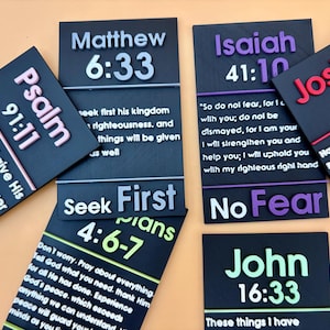 May include: A collection of rectangular black plaques featuring raised 3D lettering in various colors. Each plaque displays a specific Bible verse reference and text, including Matthew 6:33, Isaiah 41:10, Joshua 1:9, Psalm 91:11, Philippians 4:6-7, and John 16:33.