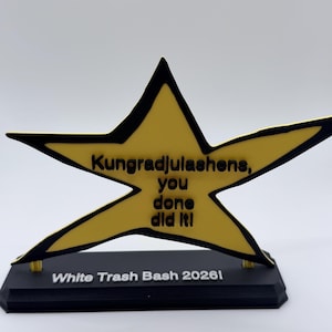 May include: A yellow star-shaped trophy with a black outline, featuring the black text "Kungradjulashens, you done did it!" on the star and "White Trash Bash 2026!" on the black rectangular base. The trophy is mounted on a black stand.