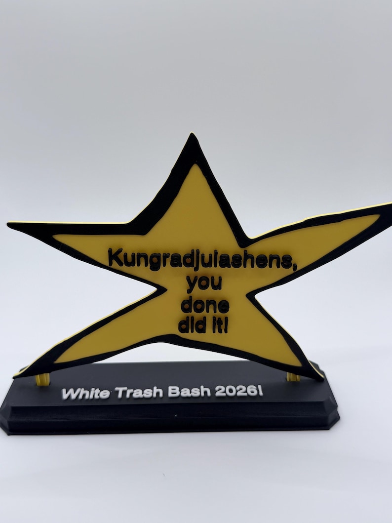 May include: A yellow star-shaped trophy with a black outline, featuring the black text "Kungradjulashens, you done did it!" on the star and "White Trash Bash 2026!" on the black rectangular base. The trophy is mounted on a black stand.