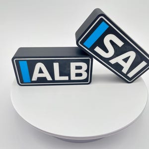以下が含まれることがあります： 白い文字と青いストライプが付いた黒い長方形のブロックが2つ。ブロックには「ALB」と「SAI」と書かれている。