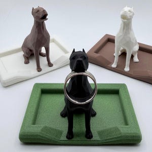 May include: A collection of dog-shaped ring holders in various colors. The black dog holds a silver ring. Other dogs are brown and white, each sitting on a matching tray. The trays are white, green, and brown.