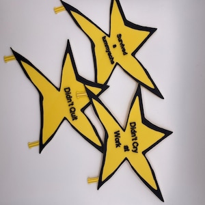 May include: Three yellow star-shaped pins with black outlines. Each star features black text: one reads 'Survived a tummyache', another 'Didn't Quit', and the third 'Didn't Cry at Work'. Each pin has a small yellow backing mechanism visible.