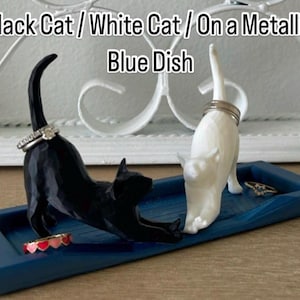 Pu&ograve; includere: Un piatto in ceramica blu con una statuetta di gatto nero e una statuetta di gatto bianco. I gatti tengono degli anelli. Il piatto &egrave; su una superficie bianca. Gatto Nero / Gatto Bianco / Su un Piatto Blu Metallico