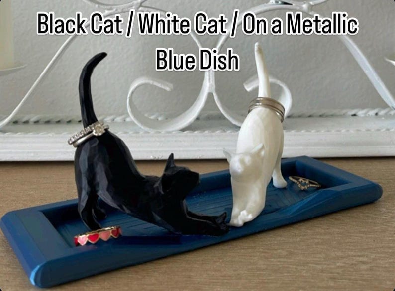 Pu&ograve; includere: Un piatto in ceramica blu con una statuetta di gatto nero e una statuetta di gatto bianco. I gatti tengono degli anelli. Il piatto &egrave; su una superficie bianca. Gatto Nero / Gatto Bianco / Su un Piatto Blu Metallico
