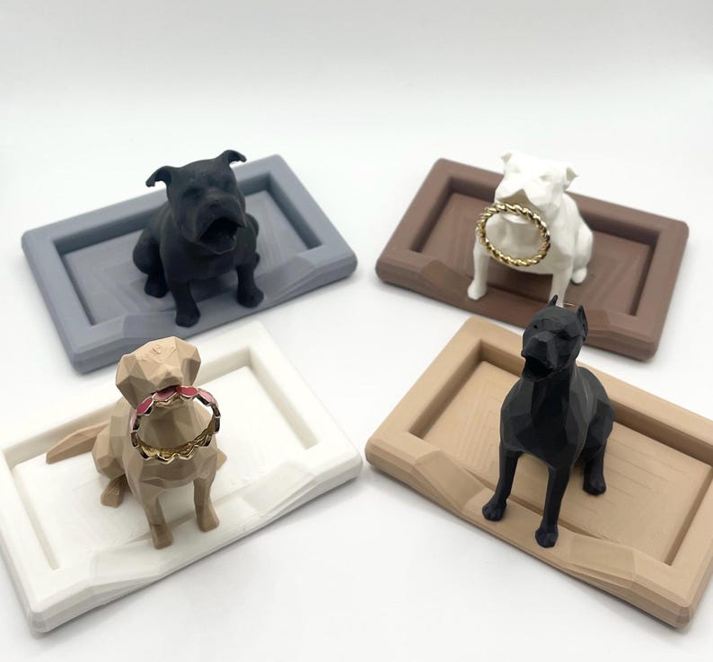 Puede incluir: Cuatro sujetadores de anillos con forma de perro en negro, blanco, beige y gris oscuro. Cada perro est&aacute; colocado en una bandeja rectangular. El perro blanco sostiene un anillo dorado, y el perro beige sostiene un anillo rojo y dorado.