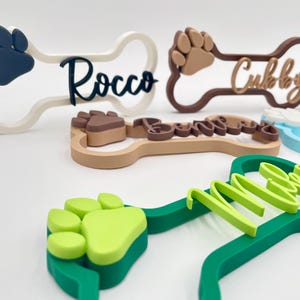 May include: A collection of bone-shaped name signs featuring raised paw prints and personalized names in various colors including white, dark blue, brown, light green, and light blue. Each sign displays a unique name such as Rocco, Cubby, and Miles.