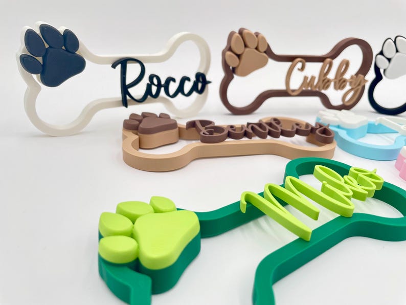 May include: A collection of bone-shaped name signs featuring raised paw prints and personalized names in various colors including white, dark blue, brown, light green, and light blue. Each sign displays a unique name such as Rocco, Cubby, and Miles.