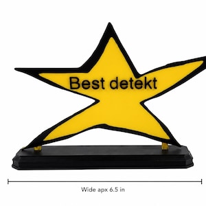 May include: A yellow star-shaped plaque with a black outline and the text "Best detekt" in black lettering. The star is mounted on a rectangular black base. The object measures approximately 4.5 inches tall and 6.5 inches wide.