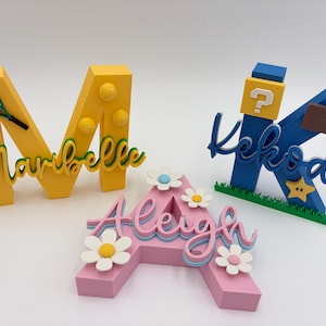 May include: Three-dimensional custom name signs. The letter "M" is yellow with tennis ball and racket accents. The letter "K" is blue with a question mark block and star. The letter "A" is pink with daisy accents.
