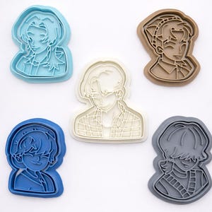 May include: Five plastic cookie cutters in various colors, including light blue, brown, white, dark blue, and gray. Each cutter features a detailed outline of a different anime-style character bust with distinct hairstyles and clothing patterns.