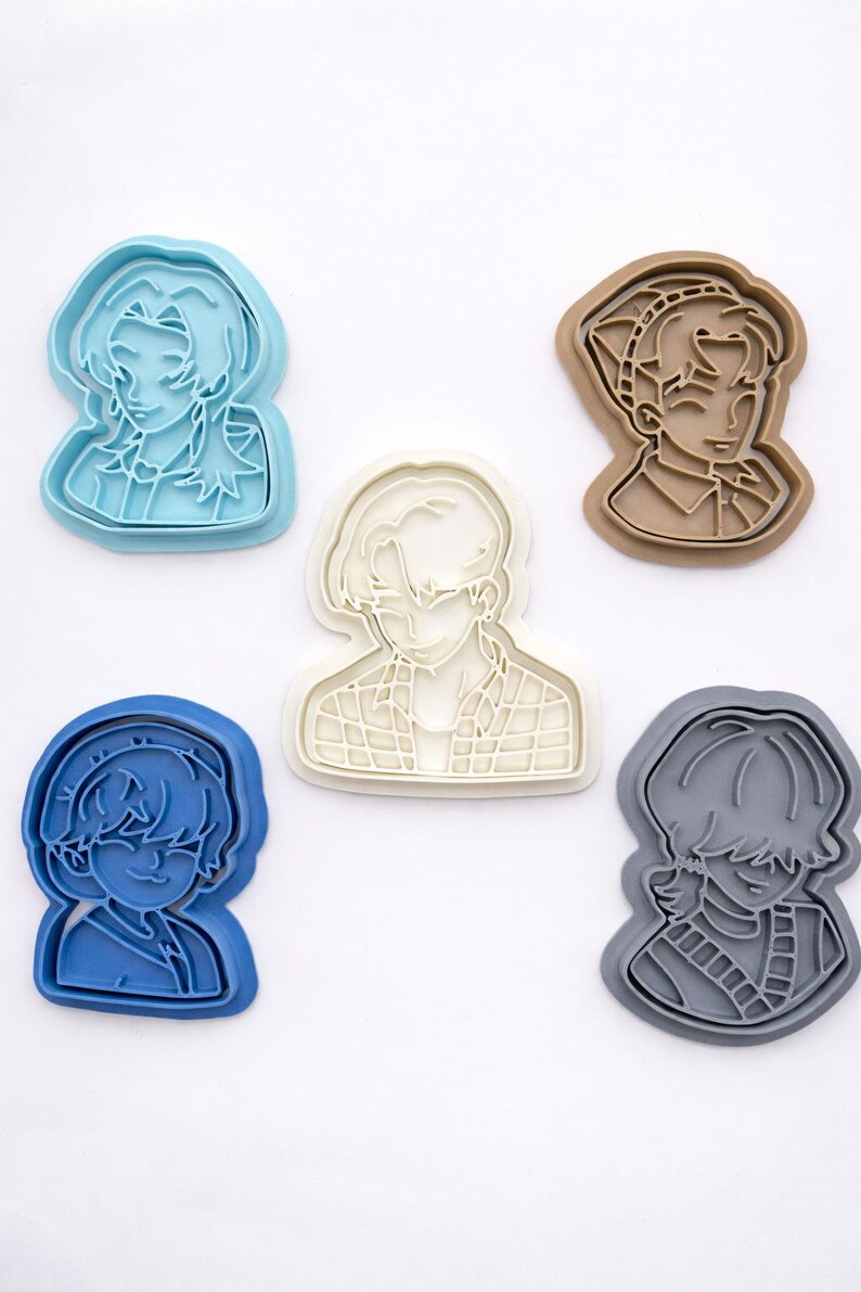 May include: Five plastic cookie cutters in various colors, including light blue, brown, white, dark blue, and gray. Each cutter features a detailed outline of a different anime-style character bust with distinct hairstyles and clothing patterns.