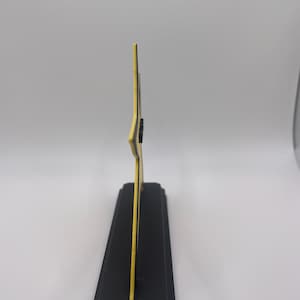 May include: A side profile view of a yellow star-shaped object mounted on a rectangular black base. The star has a thin black outline and a flat, vertical orientation. The base is solid black with beveled edges, providing a stable foundation.