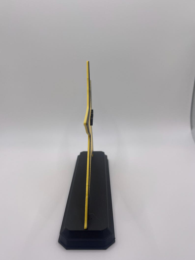 May include: A side profile view of a yellow star-shaped object mounted on a rectangular black base. The star has a thin black outline and a flat, vertical orientation. The base is solid black with beveled edges, providing a stable foundation.