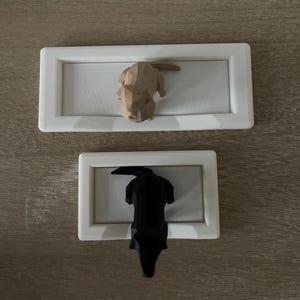 Puede incluir: Dos piezas decorativas de arte mural. La pieza superior presenta una escultura de perro geom&eacute;trico marr&oacute;n claro dentro de un marco rectangular blanco. La pieza inferior tiene una escultura de perro geom&eacute;trico negro en un marco similar.