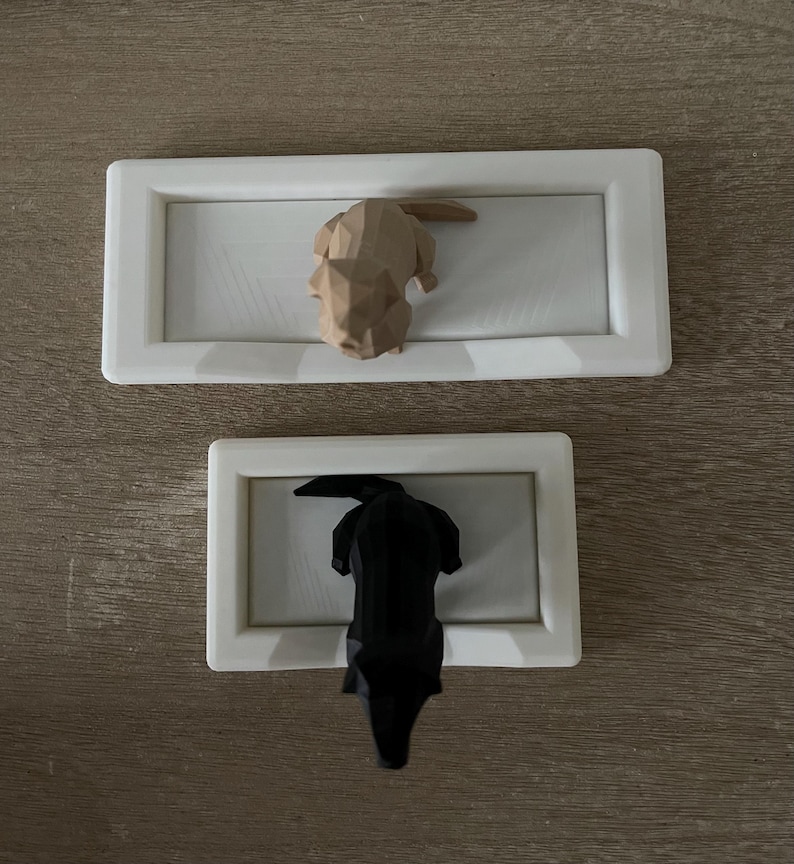Puede incluir: Dos piezas decorativas de arte mural. La pieza superior presenta una escultura de perro geom&eacute;trico marr&oacute;n claro dentro de un marco rectangular blanco. La pieza inferior tiene una escultura de perro geom&eacute;trico negro en un marco similar.