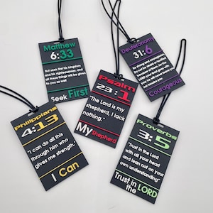 May include: A collection of black rectangular tags with Bible verses and references. Each tag features a different verse in white, green, red, purple, and gold lettering. The tags are attached to black cords.