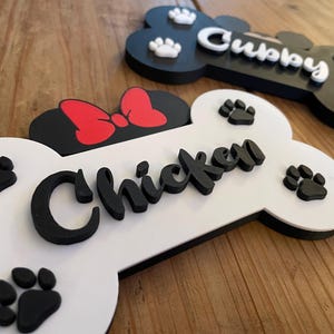 May include: Bone-shaped pet name signs in black and white with paw prints. One sign has the name "Chicken" in black script, and the other has "Cubby" in white script. A red bow is on the "Chicken" sign.