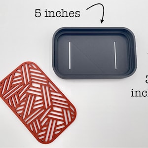 May include: A dark gray rectangular container, 5 inches long and 3 inches wide, with two white lines and a diagonal line. A red, geometric patterned mat is next to the container.