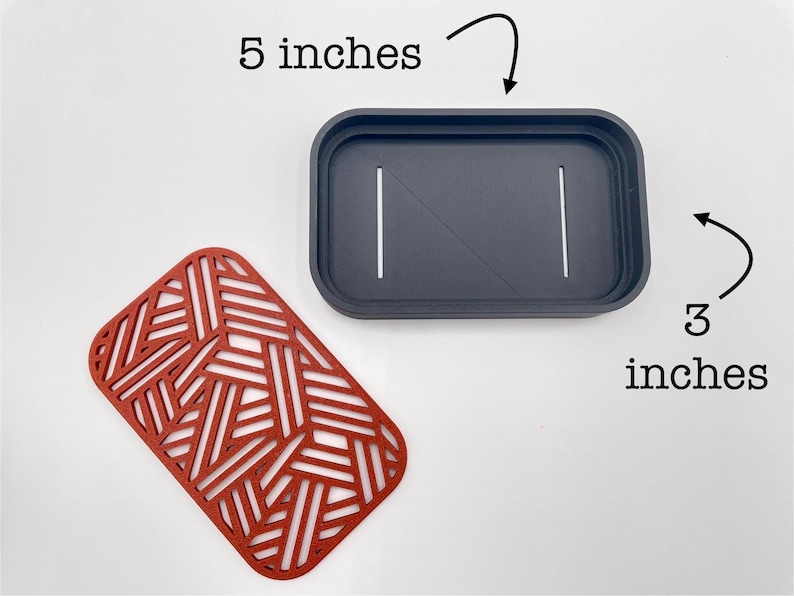 May include: A dark gray rectangular container, 5 inches long and 3 inches wide, with two white lines and a diagonal line. A red, geometric patterned mat is next to the container.
