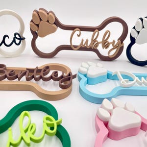 May include: A collection of bone-shaped name signs in various colors including white, brown, blue, green, and pink. Each sign features a raised paw print and a custom name in cursive script, such as Rocco, Cubby, Bentley, and Miko, made of plastic.