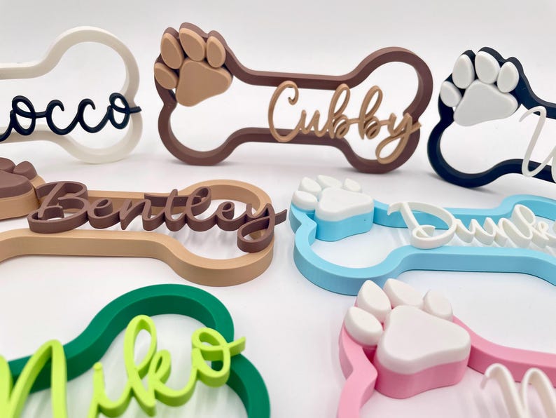 May include: A collection of bone-shaped name signs in various colors including white, brown, blue, green, and pink. Each sign features a raised paw print and a custom name in cursive script, such as Rocco, Cubby, Bentley, and Miko, made of plastic.