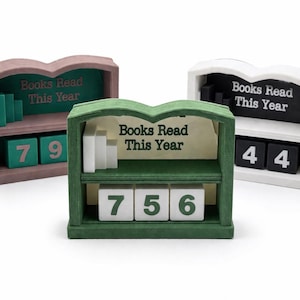 May include: Three wooden book counters shaped like bookshelves in pink, green, and white. Each has a shelf with small book-shaped blocks and number blocks to track books read. The text "Books Read This Year" is displayed.