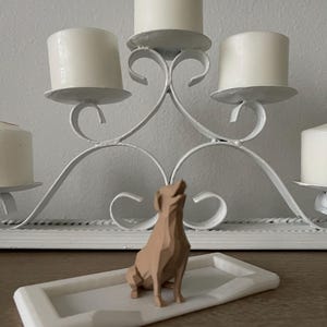 Puede incluir: Un candelabro de metal blanco con cinco velas blancas. Una figura geom&eacute;trica de perro de color marr&oacute;n claro se encuentra sobre una bandeja rectangular blanca frente al candelabro. El perro est&aacute; sentado con la cabeza levantada.
