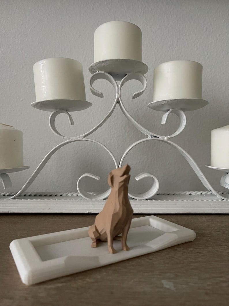 Puede incluir: Un candelabro de metal blanco con cinco velas blancas. Una figura geom&eacute;trica de perro de color marr&oacute;n claro se encuentra sobre una bandeja rectangular blanca frente al candelabro. El perro est&aacute; sentado con la cabeza levantada.