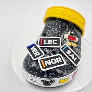 Peut inclure: Sabot Crocs noir avec une bride jaune, orné de quatre breloques rectangulaires avec les inscriptions "VER", "LEC", "NOR" et "SAI". Un charm Mickey Mouse et un motif blanc décorent la chaussure.