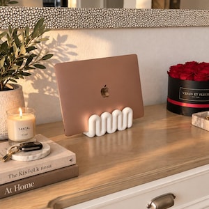 May include: A white, wavy, sculptural laptop stand holding a rose gold laptop. The stand features a continuous, undulating, serpentine design that creates multiple slots for upright storage, providing a stable base for the electronic device.