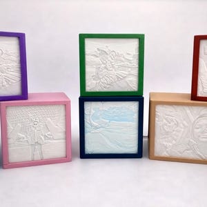May include: A collection of square, framed photo blocks in various colors, including purple, pink, green, and red. Each block features a white, 3D-printed image of a photograph. The frames are square and the images are in relief.