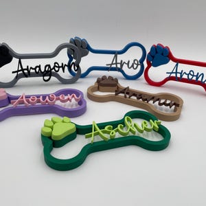 May include: A collection of six 3D-printed bone-shaped name signs in various colors, including grey, blue, red, purple, brown, and green. Each sign features a raised paw print and a custom name in cursive script, such as Aragorn, Ario, Arondir, Aowen, Arawn, and Ascher.