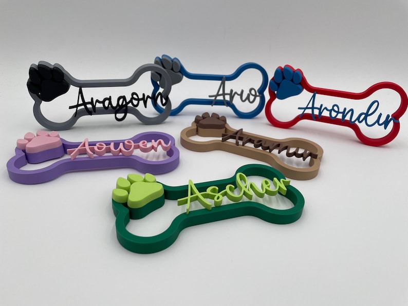 May include: A collection of six 3D-printed bone-shaped name signs in various colors, including grey, blue, red, purple, brown, and green. Each sign features a raised paw print and a custom name in cursive script, such as Aragorn, Ario, Arondir, Aowen, Arawn, and Ascher.