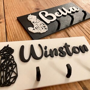 May include: Two white wooden key hooks with black geometric cat silhouettes and black lettering. One hook says "Bella" and the other says "Winston".