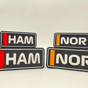 以下が含まれることがあります： 白文字の黒い長方形のラベルが2組。上のラベルは「HAM」と「NOR」で、それぞれ赤とオレンジのストライプが入っている。下のラベルは「HAM」と「NOR」で、それぞれ赤とオレンジのストライプが入っている。