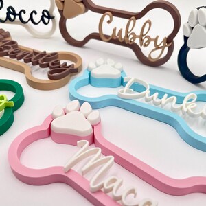 May include: A collection of custom 3D-printed dog bone-shaped name tags in various colors including pink, blue, green, brown, and black. Each tag features a raised paw print accent and a personalized name in a cursive script font on the front.