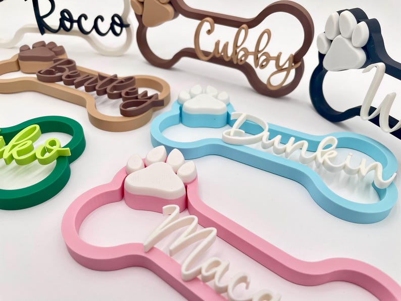 May include: A collection of custom 3D-printed dog bone-shaped name tags in various colors including pink, blue, green, brown, and black. Each tag features a raised paw print accent and a personalized name in a cursive script font on the front.