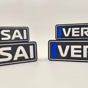 以下が含まれることがあります： 青いアクセントが付いた黒と白の長方形のステッカーが2セット。各セットの上のステッカーには「VER」、下のステッカーには「ISAI」と書いてあります。