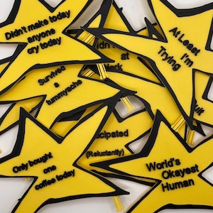 May include: A collection of yellow star-shaped badges with black outlines and black text. The phrases include 'Didn't make anyone cry today', 'At Least I'm Trying', 'Survived a tummyache', 'Only bought one coffee today', 'Participated (Reluctantly)', and 'World's Okayest Human'.