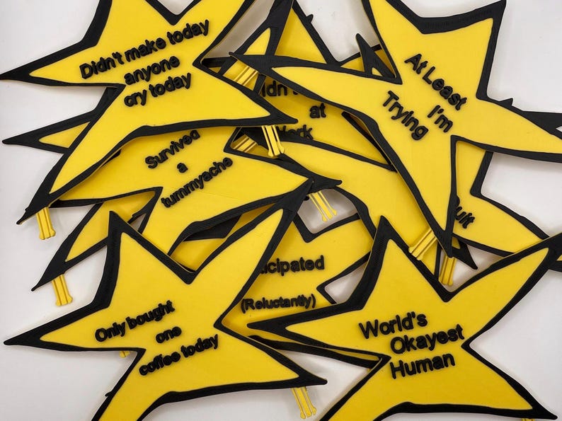 May include: A collection of yellow star-shaped badges with black outlines and black text. The phrases include 'Didn't make anyone cry today', 'At Least I'm Trying', 'Survived a tummyache', 'Only bought one coffee today', 'Participated (Reluctantly)', and 'World's Okayest Human'.