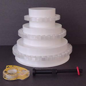 May include: A white cake stand with five tiers, each with a decorative, clear plastic trim. The stand is on a gray surface. A black tool with a red ring is in the foreground, along with a roll of clear tape.